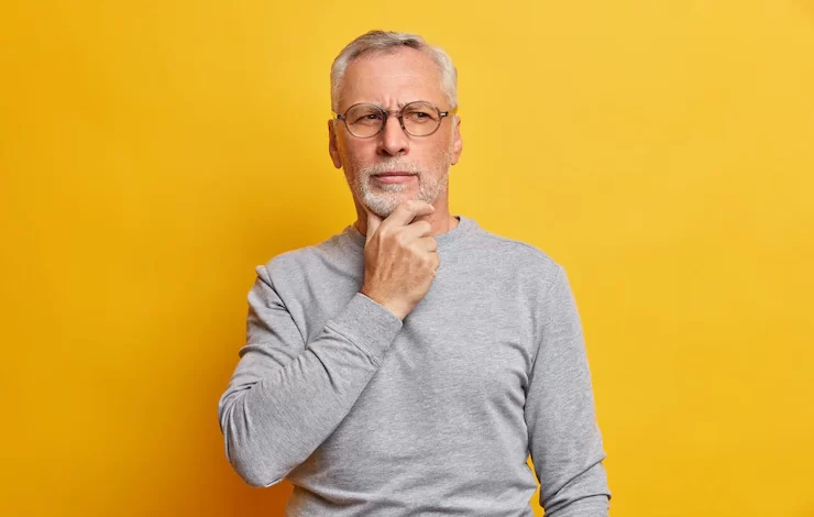 senior thougthful man holds chin looks pensively aside makes plannings wears spectacles casual grey jumper isolated vivid yellow wall 273609 44143